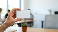 Male hand holding blank white business card in modern office environment with colleagues working in background, suitable for corporate branding and professional networking materials.
