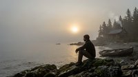 Silhouette of contemplative man sitting on rocky shoreline at misty sunrise with wooden boats and cabin among pine trees creating peaceful lakeside scene.