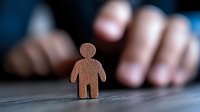 Wooden human figure silhouette standing alone on dark surface with blurred hands in background, representing solitude, personal growth, and human relationships.
