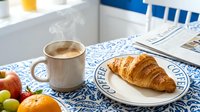 Steaming coffee mug and fresh croissant on a blue floral tablecloth with fruit and newspaper, cozy morning breakfast scene.