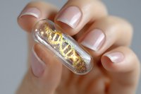 Transparent capsule filled with golden glitter held between manicured fingers with natural pink nail polish against blurred background. Medical concept.