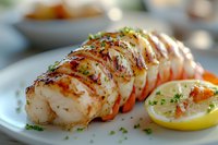 Grilled lobster tail with butter sauce and fresh herbs garnished with lemon slice on white plate, close up view showing juicy meat texture and caramelized surface.