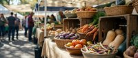 Farmers market produce stand displaying fresh vegetables including carrots, squash, potatoes and root vegetables in wicker baskets with blurred shoppers browsing in background.