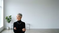 Senior woman with short gray hair wearing black turtleneck, arms crossed, standing in minimalist office space with plant and window light.