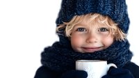 Young child wearing navy blue knitted winter hat and scarf holds white ceramic mug, smiling warmly against white background. Cozy winter lifestyle portrait.