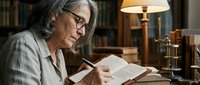 Senior woman with gray hair and glasses reading book in cozy library setting with warm lamp light. Academic research and lifelong learning concept.