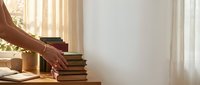 Hand reaching for stack of books on wooden table near bright window with warm sunlight streaming through sheer curtains in cozy reading space.