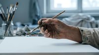 Artist hand holding paintbrush in creative studio workspace with art supplies and natural light for painting and drawing projects.