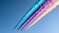 Colorful airplane contrails streak across bright blue sky creating vibrant parallel lines in turquoise, navy blue, pink and orange colors against clear atmosphere.