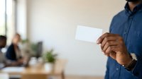 Male professional of South Asian descent wearing a navy blue button-up shirt holding a blank white business card in an office environment with colleagues and plants in background.