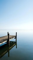 Wooden dock extending into calm, mirror-like water under clear blue sky. Weathered pier with dark posts creates serene, minimalist landscape. Ideal for travel, nature, and wellness concepts.
