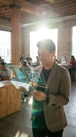 Young Asian businessman using holographic tablet technology in modern coworking office space with natural sunlight streaming through windows.