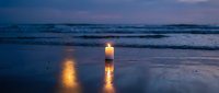 Single candle glowing on wet beach sand at twilight with ocean waves and purple sky creating peaceful meditation atmosphere for wellness and spirituality concepts.