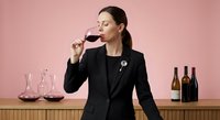 Professional female sommelier in black suit tasting red wine at elegant bar counter with wine bottles and glasses on pink background for hospitality industry.