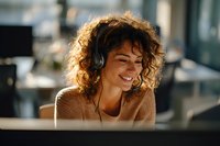 Young woman with curly hair wearing headphones smiles while working on computer in bright modern office space.