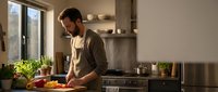 Young bearded man in apron preparing fresh vegetables in modern kitchen with natural lighting and herbs on counter for healthy home cooking.