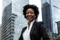 Confident African American businesswoman with natural hair smiling against modern urban backdrop of glass skyscrapers, wearing professional black suit and white shirt.