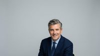 Middle-aged man with gray hair and light skin tone wearing dark navy blazer and blue tie against neutral gray background for corporate portraits.