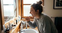 Young woman brushing teeth in modern bathroom with natural light from window. Daily morning hygiene routine and oral care habits for healthy lifestyle.