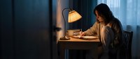 Young woman writing in notebook at wooden desk with warm lamp light during evening hours creating cozy study atmosphere for productivity and focus.