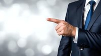 Businessman in navy suit with blue tie pointing finger to side against blurred silver bokeh background, conveying direction or choice in professional setting.
