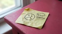 Crumpled yellow sticky note with sad face drawing and service text on pink surface near window expressing customer dissatisfaction feedback concept.