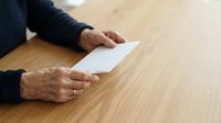 Mature man with light skin tone holding blank white envelope on wooden table surface, suitable for business correspondence and communication concepts.