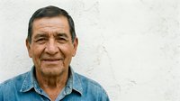 Senior man with dark hair and warm smile wearing denim shirt, portrait against white textured wall background. Suitable for healthcare, family, community and lifestyle editorial use.