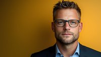 Confident caucasian businessman wearing glasses and navy suit jacket poses against vibrant yellow background, professional corporate headshot with modern aesthetic appeal.