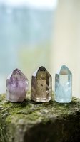 Three healing crystal points in purple amethyst, smoky quartz and blue aquamarine arranged on moss-covered stone surface for spiritual wellness and meditation.