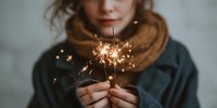 Young woman holding burning sparkler creating magical golden sparks and light trails during winter celebration or holiday season.