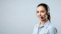 Professional female customer service representative with headset smiling at camera wearing light blue shirt against neutral background for business communications.