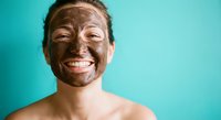 Young Caucasian woman with chocolate face mask smiling at camera against turquoise background. Natural skincare and beauty treatment concept for spa wellness.