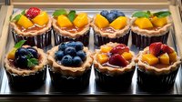 Fresh fruit mini tarts with blueberries, strawberries, and mango pieces arranged in crispy pastry shells on display in bakery case, dessert catering.
