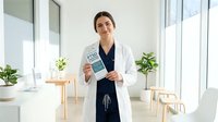 Young Hispanic female doctor in white coat holding medical brochure in modern bright clinic office with plants and natural lighting for healthcare marketing.
