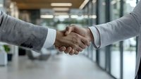 Close-up of business handshake between two professionals in gray suits against blurred modern office background, symbolizing partnership and agreement.