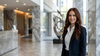 Professional businesswoman with long brown hair in navy blazer and white shirt smiling confidently in modern corporate office lobby with marble columns.