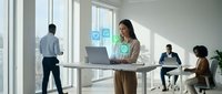 Woman with long dark hair, South Asian ethnicity, wearing casual business attire, working at standing desk with laptop in modern office environment with social media icons displayed, colleagues collaborating in background.
