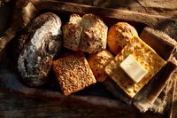 Assortment of artisanal bread loaves and rolls with butter on rustic wooden surface, showcasing variety of textures and crusts in warm lighting.