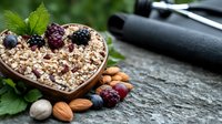 Heart shaped bowl filled with granola, topped with fresh berries and surrounded by nuts on dark rustic wooden surface. Healthy breakfast concept with natural ingredients.