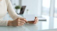Cropped view of woman holding blank white business card at desk with laptop and plant in blurred office background for corporate branding.
