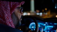 Middle Eastern man in traditional keffiyeh headdress driving car at night, illuminated by blue dashboard lights, profile view showing focused expression and glasses.