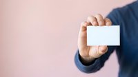 Male professional in dark blue business attire holding blank white business card against pink background, ready for contact information or branding mockup.