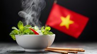 Steaming hot Asian noodle soup in white ceramic bowl with fresh herbs, chopsticks and Vietnamese flag in background, dramatic food photography setup.