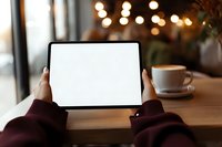 Person holding blank tablet screen in cozy cafe with coffee cup on wooden table and soft bokeh lights in background.