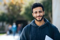 Young Middle Eastern male student smiling outdoors on university campus wearing dark hoodie and carrying notebook, with blurred people in background.