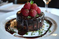 Decadent chocolate cake topped with fresh raspberries and dark chocolate ganache, garnished with mint leaves on white plate with elegant sauce presentation.