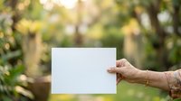 Mature woman holding blank white card outdoors in garden setting with blurred green foliage background, suitable for advertising and marketing campaigns.