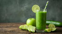 Fresh green smoothie with lime slice and striped straw surrounded by green apple, cucumber and lime wedges on rustic wooden table for healthy lifestyle.