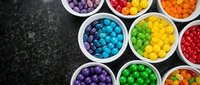 Colorful candy balls sorted by vibrant hues in white bowls on dark textured surface for confectionery and sweet treat designs.